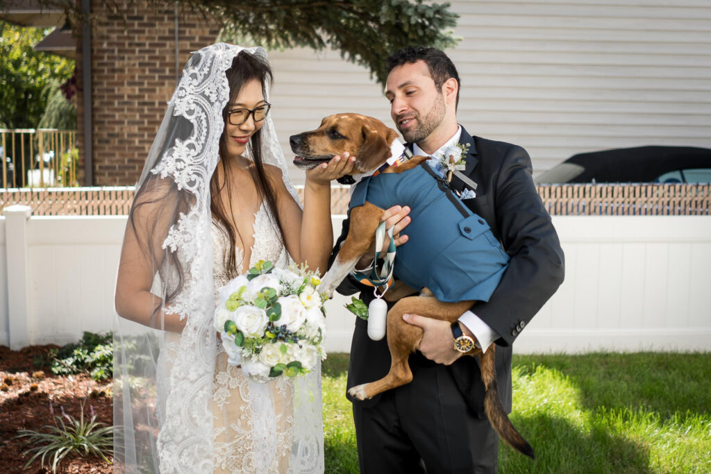 Newlyweds Ruby and Rob holding their dog GriGri in blue suit jacket during Fair Lawn backyard home wedding portraits