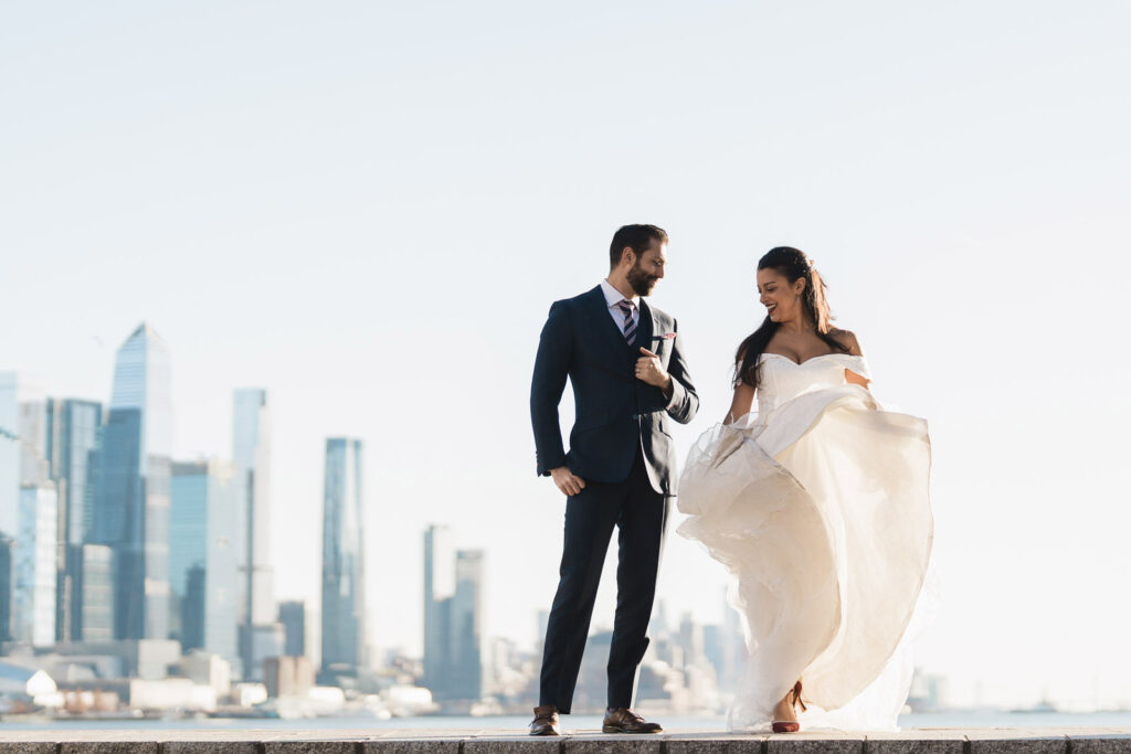Joanna's lace wedding dress flowing in wind with Alex on waterfront NYC skyline Port Imperial romantic photos