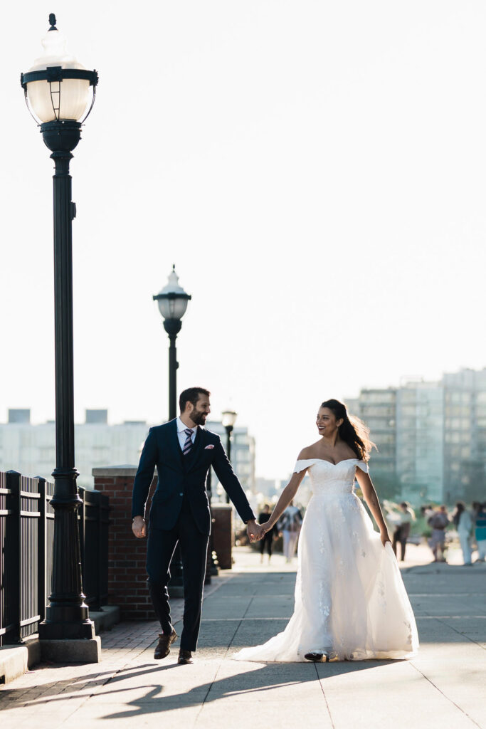 Joanna and Alex holding hands walking on Port Imperial promenade with lamppost and Manhattan skyline West New York NJ wedding photographer
