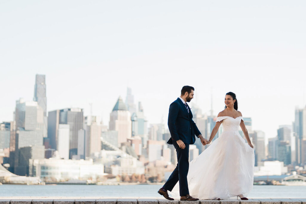 Joanna and Alex walking hand in hand on Port Imperial promenade with NYC skyline and One World Trade Center West New York