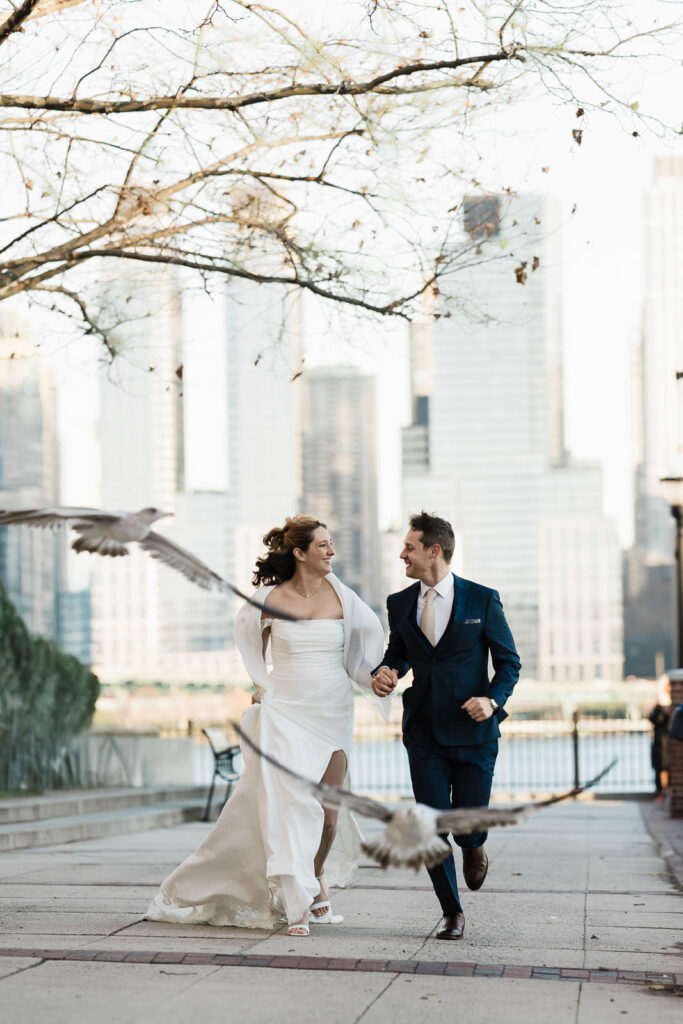 Luz and Stephan running with birds and Manhattan skyline playful wedding photography Port Imperial West New York
