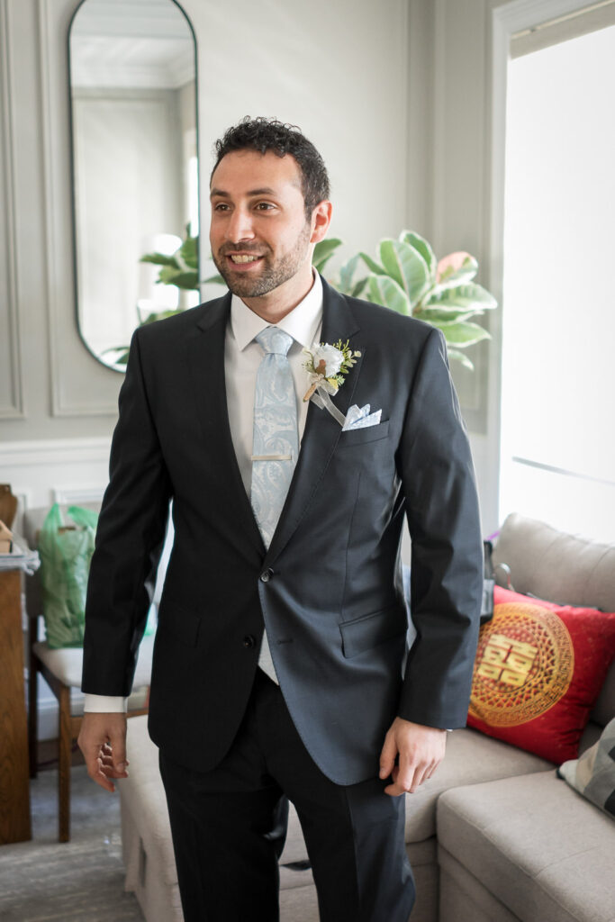 Groom Rob smiling in charcoal suit with light blue tie and boutonniere during Fair Lawn NJ home wedding getting ready