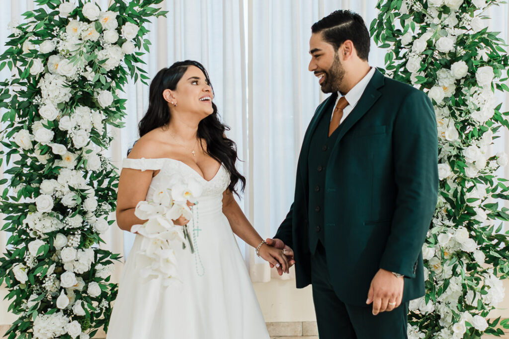 Bride and groom laughing together in front of white floral arch at northern New Jersey wedding