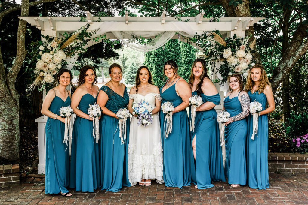 Full bridal party with bridesmaids in teal gowns and bride under decorated white pergola at Smithville Inn lakeside ceremony NJ
