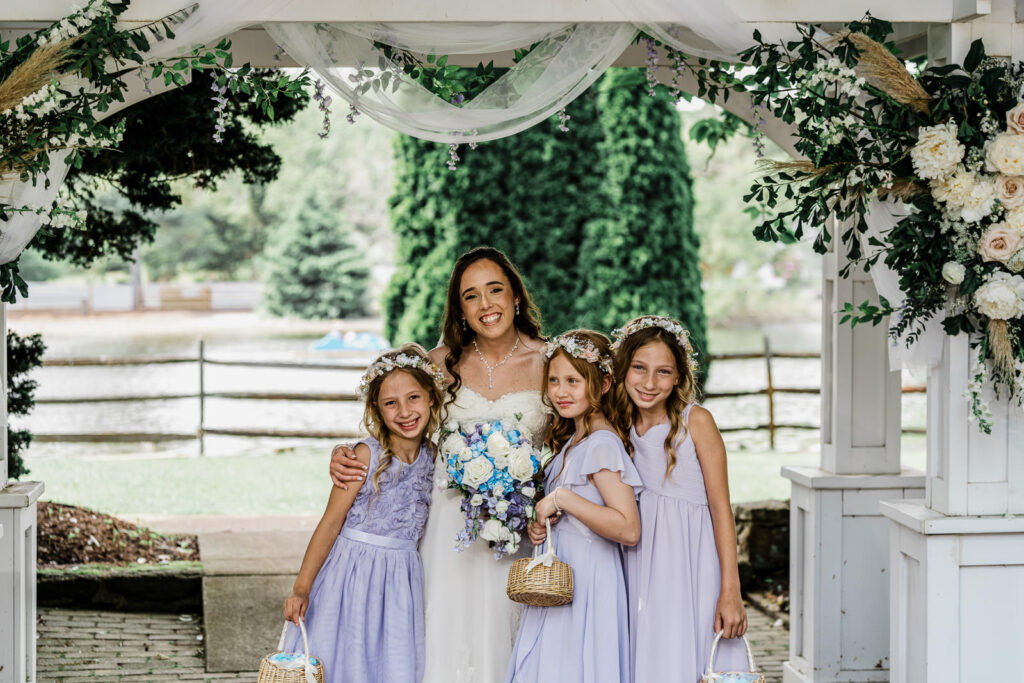 Bride with three young flower girls in purple dresses under white floral pergola at Smithville Inn ceremony in New Jersey