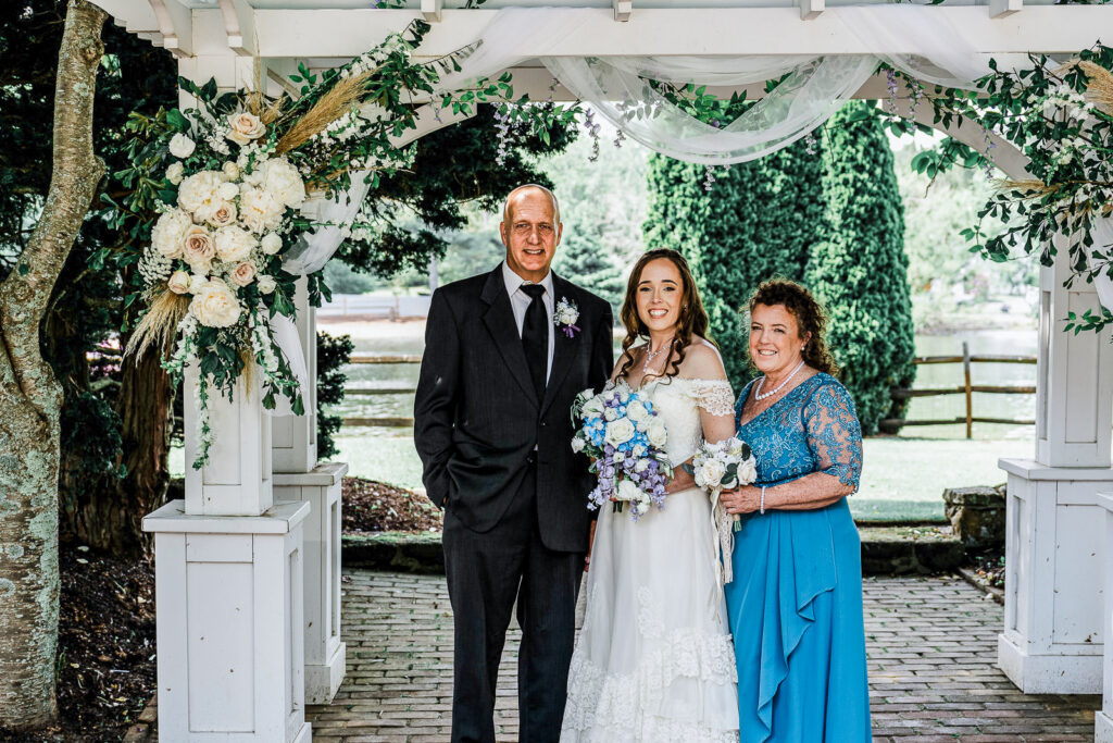Bride standing with parents under white floral pergola at lakeside ceremony altar at Smithville Inn in Absecon New Jersey