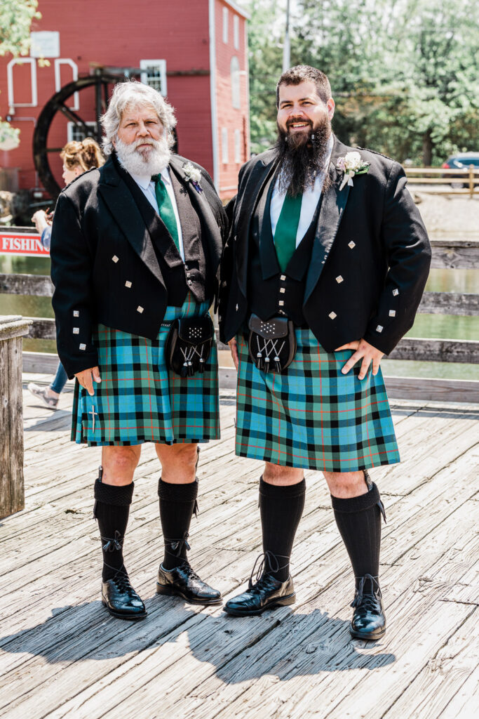 Scottish groom and father wearing traditional tartan kilts standing together at Smithville Inn with red barn backdrop in New Jersey wedding