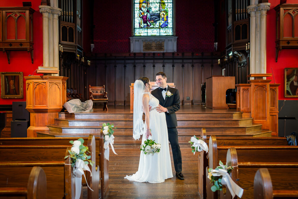 Bride and groom standing in Kirkpatrick Chapel center aisle with dramatic red uplighting and stained glass window, Alex Kaplan Photography New Brunswick