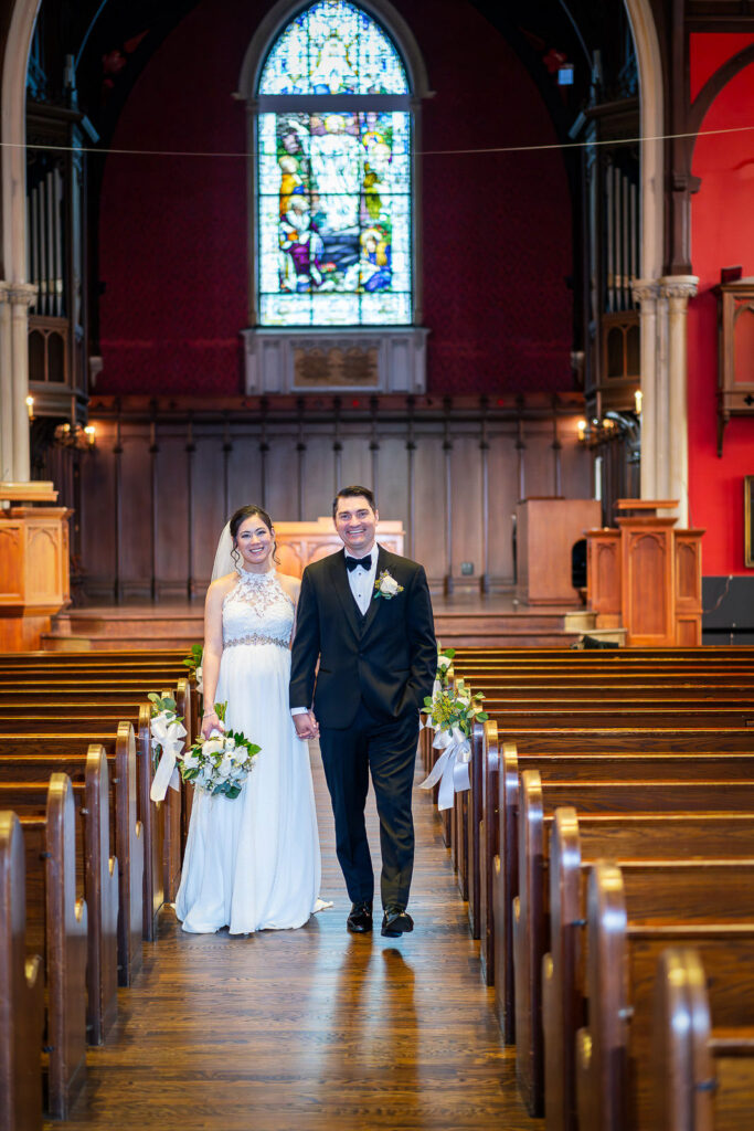 Bride and groom walking hand in hand down Kirkpatrick Chapel aisle with dramatic stained glass window and red accent lighting, Alex Kaplan Photography