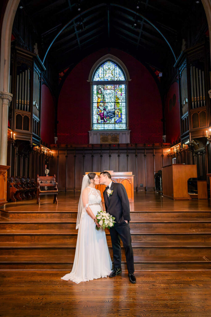 Bride and groom first kiss at Kirkpatrick Chapel wedding altar with dramatic burgundy walls and Gothic stained glass, photographed by Alex Kaplan