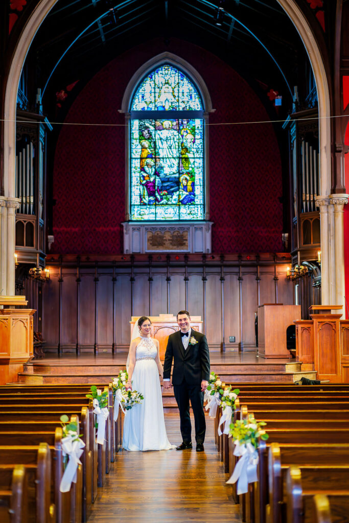 Wide angle wedding portrait down Kirkpatrick Chapel aisle showing vaulted ceilings, stained glass, and Gothic architecture, New Brunswick photography by Alex Kaplan