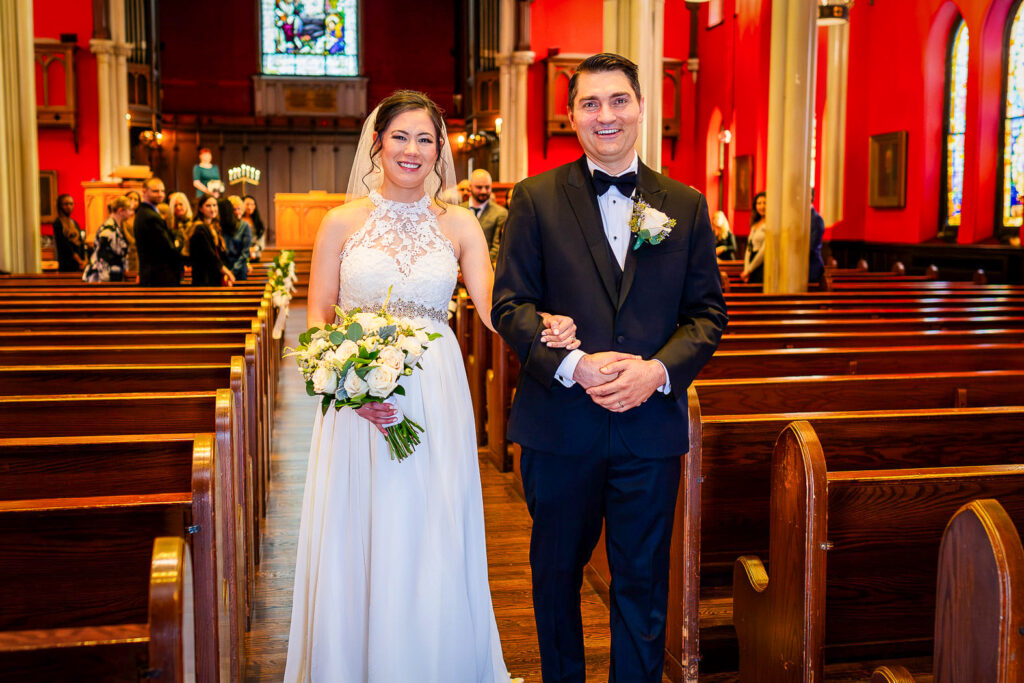 Newly married couple standing in chapel aisle at Kirkpatrick Chapel with red accent lighting and stained glass window, New Brunswick wedding by Alex Kaplan
