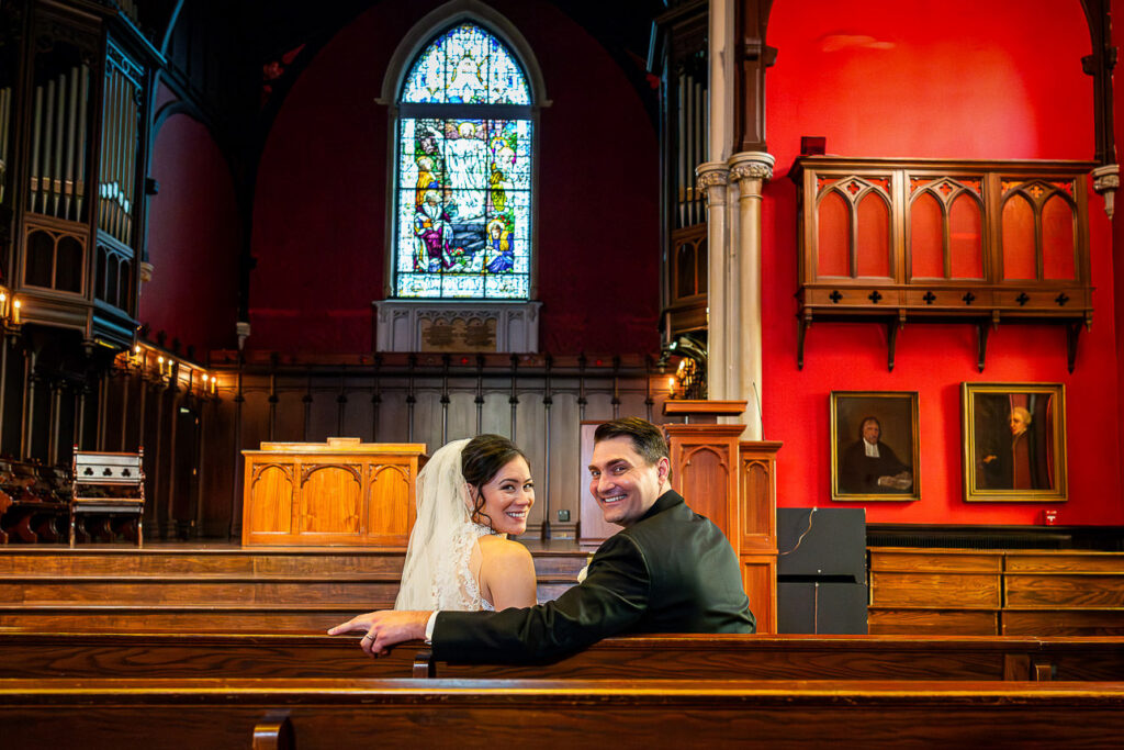 Intimate moment of bride and groom seated in chapel pew at Kirkpatrick Chapel with stained glass window, New Brunswick wedding photography by Alex Kaplan