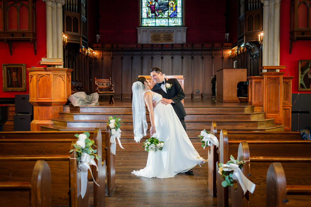 Romantic dip pose of wedding couple in Kirkpatrick Chapel aisle with red walls and white floral pew decorations, photographed by Alex Kaplan