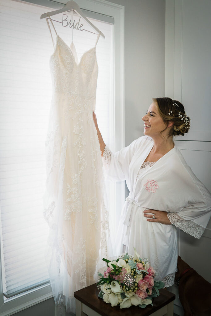 Bride in white robe smiling at her lace wedding gown hanging on personalized hanger at Tides Estate