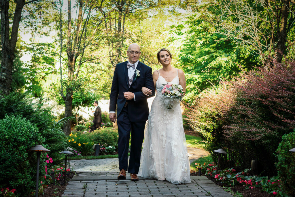 Father escorting bride down brick garden path surrounded by colorful flowers and lush greenery at Tides Estate