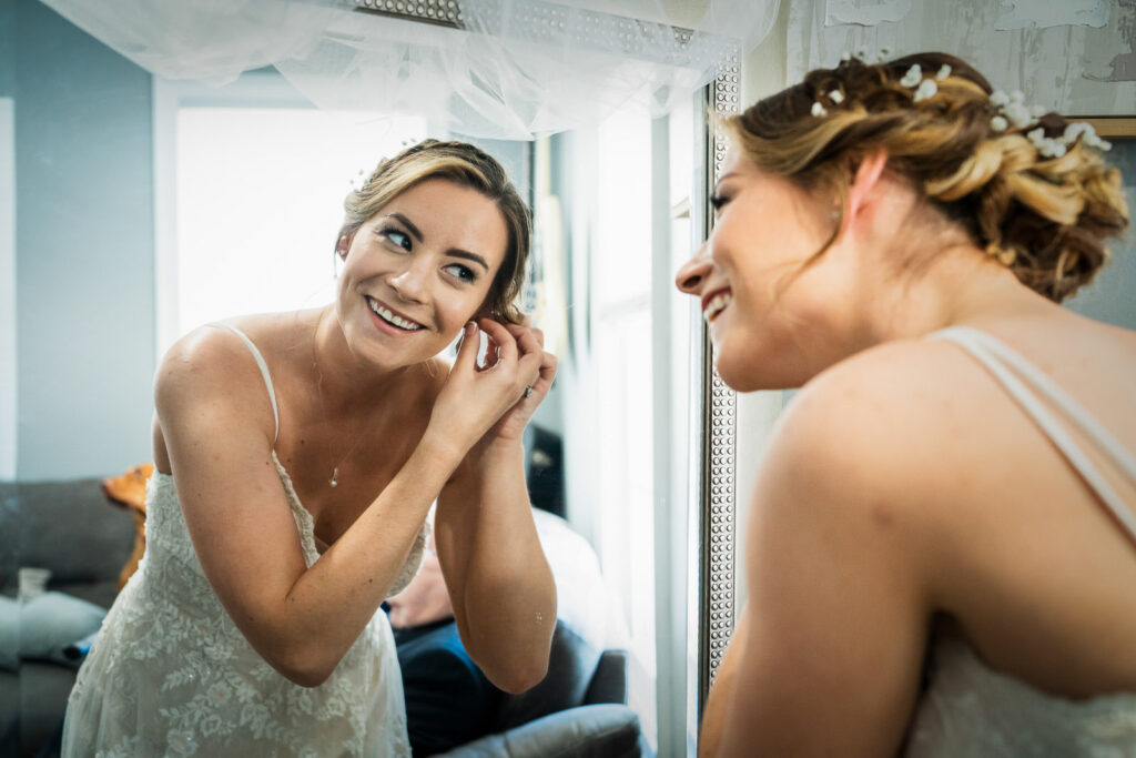 Bride putting on pearl earrings while looking in ornate mirror during Tides Estate wedding preparation