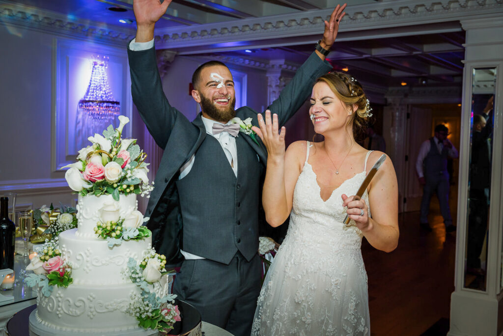 Groom celebrating with arms raised while bride laughs during cake cutting at Tides Estate reception