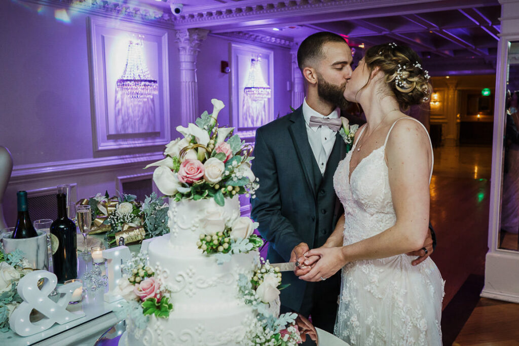 Couple kissing during cake cutting with elegant three-tier wedding cake decorated with fresh flowers at Tides Estate
