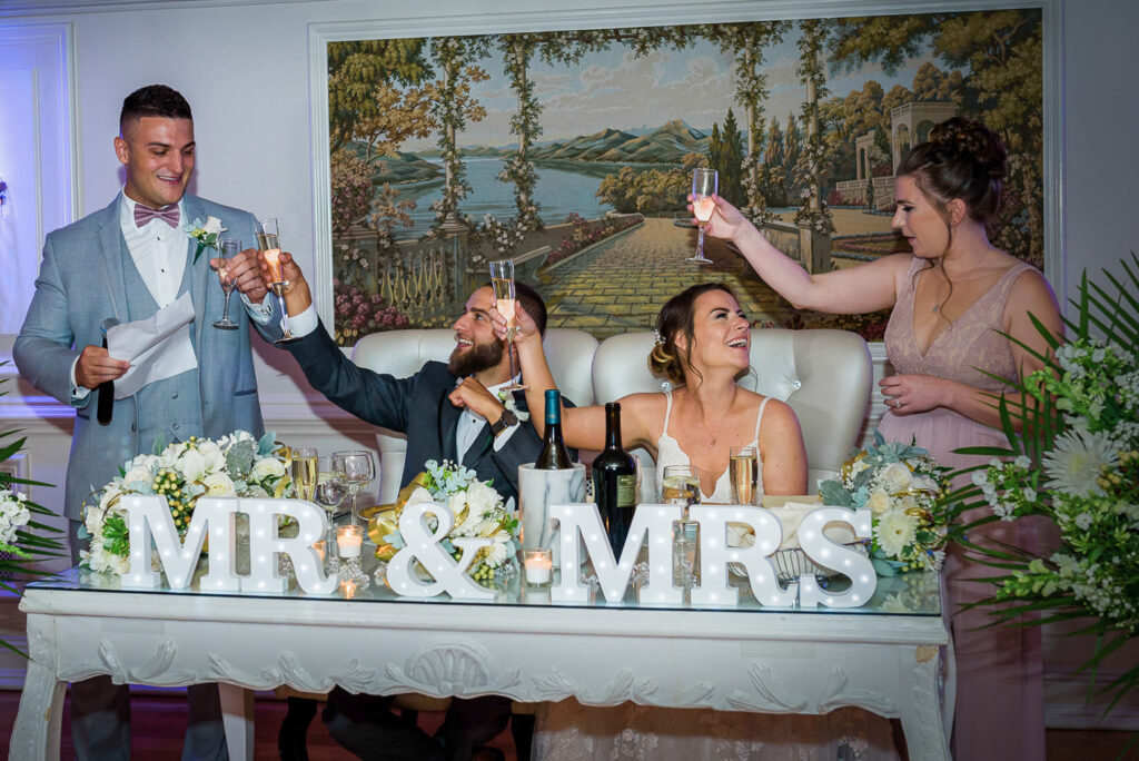 Wedding party raising champagne glasses in celebratory toast at head table with Italian landscape mural