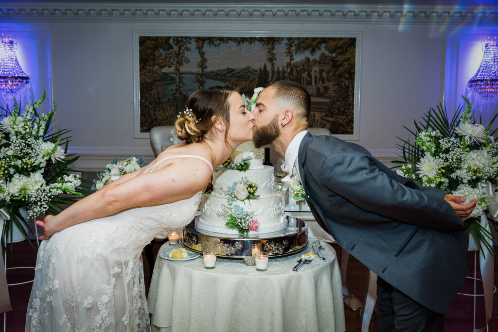 Bride and groom leaning across wedding cake table for romantic kiss in Tides Estate ballroom with Italian mural backdrop