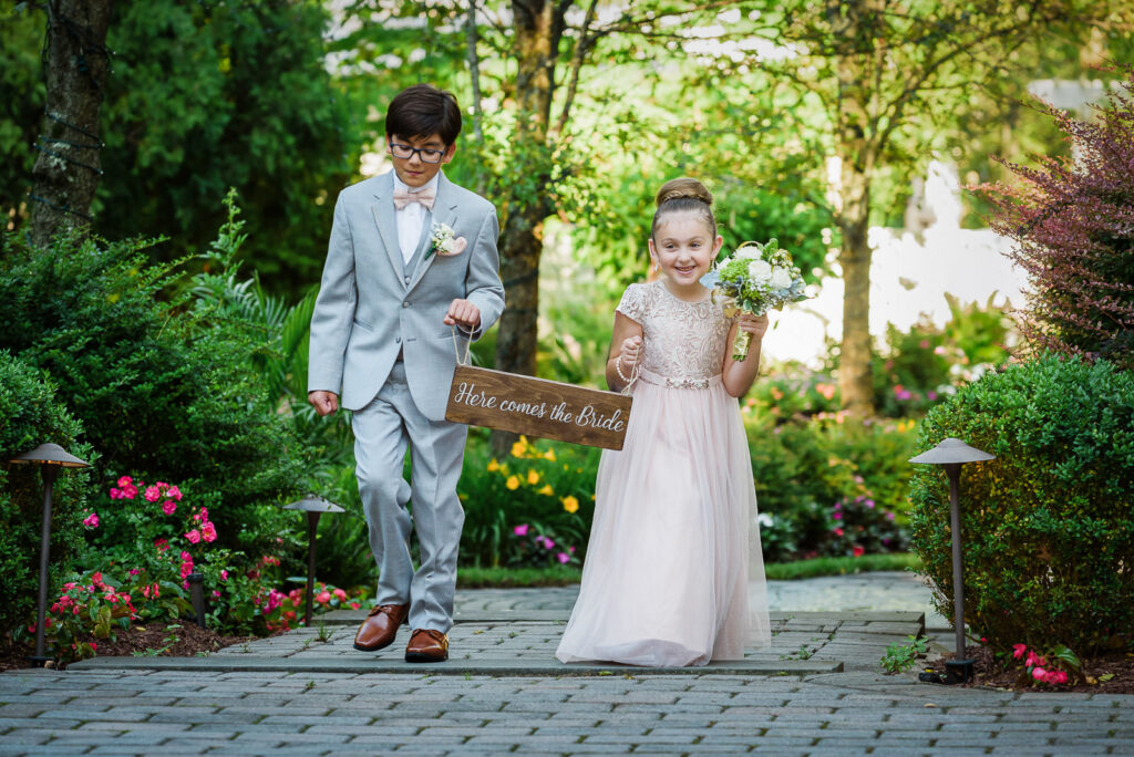 Flower girl in pink tulle dress and ring bearer in gray suit walking brick path through blooming gardens at Tides Estate