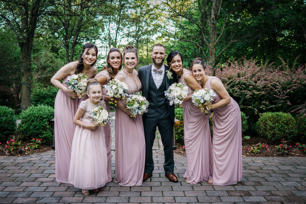 Groom surrounded by bridesmaids in blush pink gowns and flower girl at Tides Estate