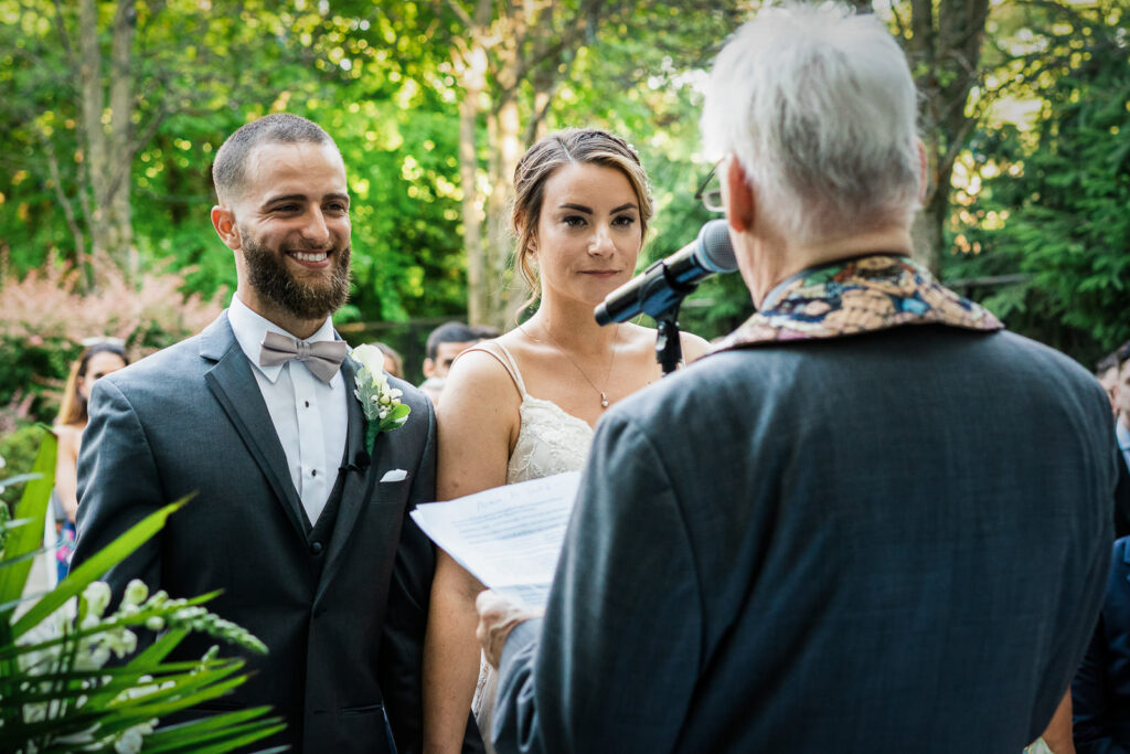 Groom smiling at bride during garden ceremony with officiant in floral vest at Tides Estate