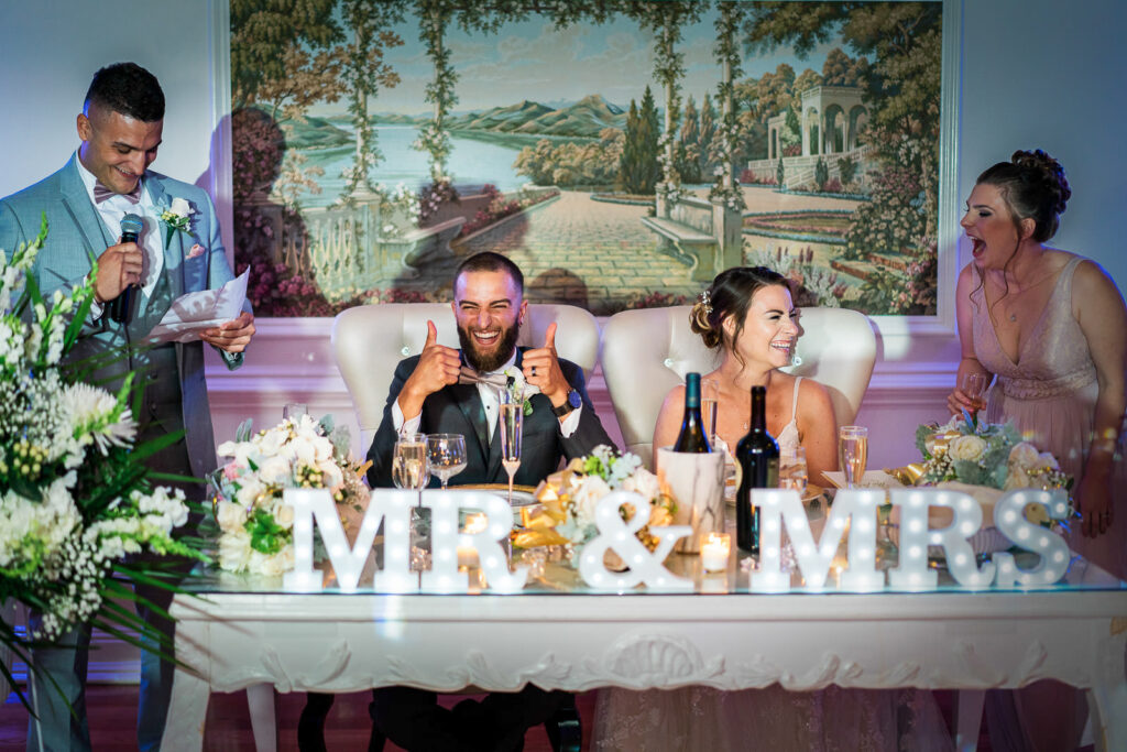 Best man giving toast while bride and groom react with laughter at sweetheart table with scenic mural backdrop