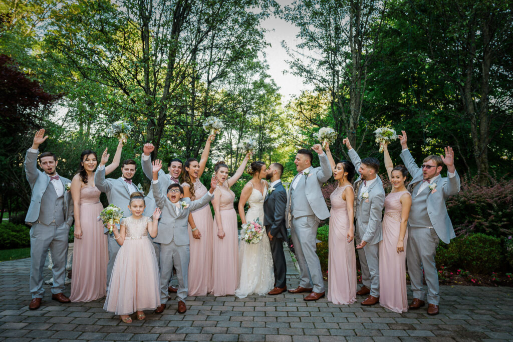 Full wedding party with raised arms celebrating with couple kissing in center at Tides Estate gardens