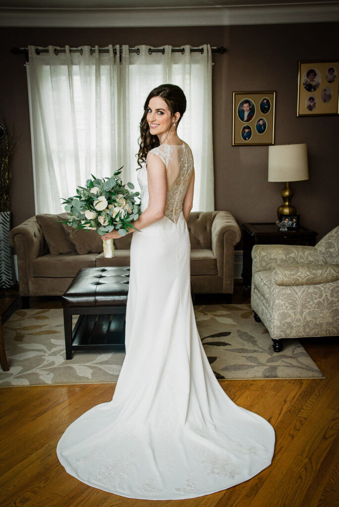 Bride in elegant lace wedding dress with illusion back holding eucalyptus and white rose bouquet before Our Lady of Hope Church ceremony