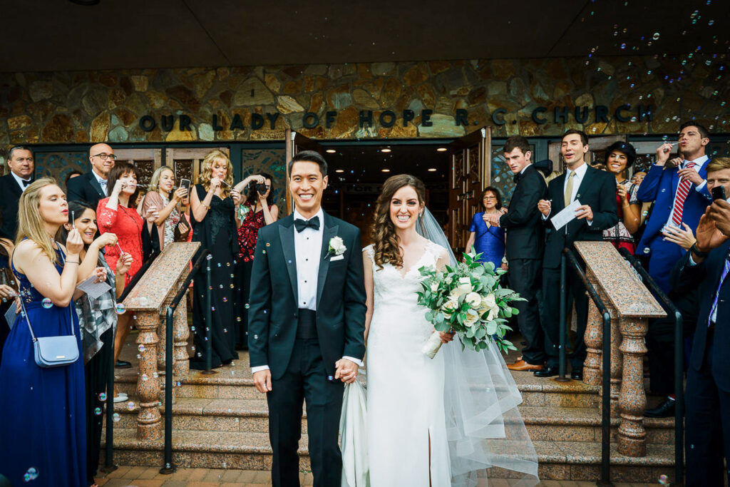 Bride in cathedral veil and groom surrounded by bubbles during church exit celebration at Glen Cove wedding