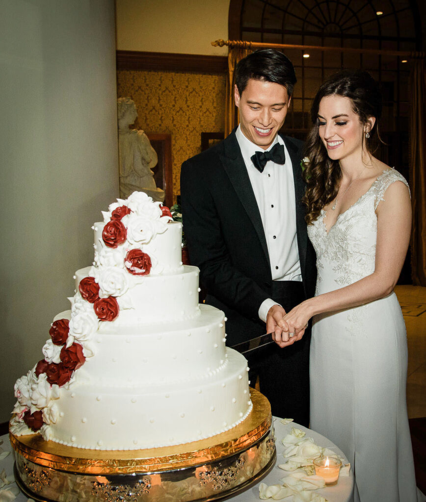 Bride and groom cutting four-tier white wedding cake with red and white sugar flowers at Metropolitan Caterers Glen Cove