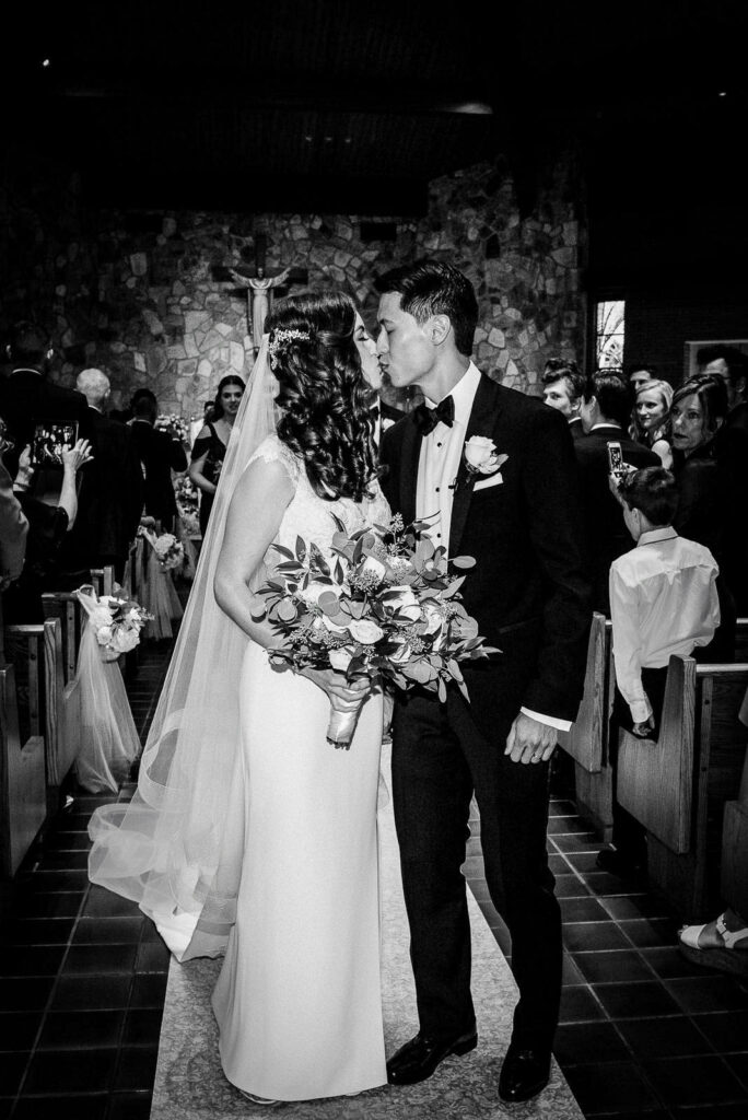 Black and white photo of newlyweds first kiss in church aisle after wedding ceremony at Our Lady of Hope Church