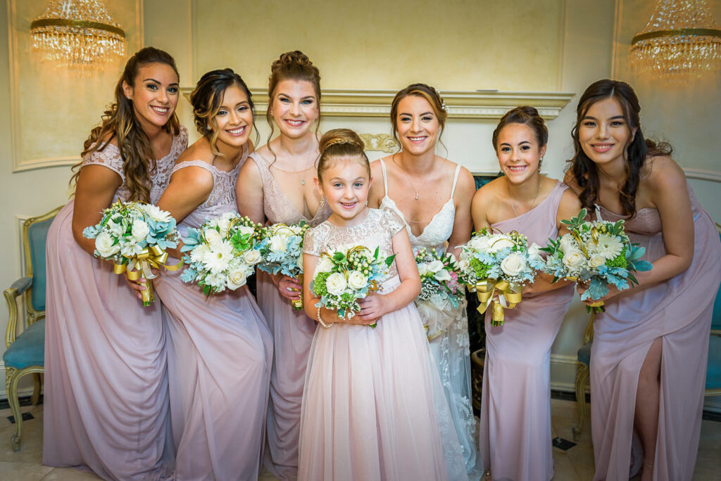 Bridesmaids in blush pink gowns with flower girl posing under crystal chandeliers at Tides Estate