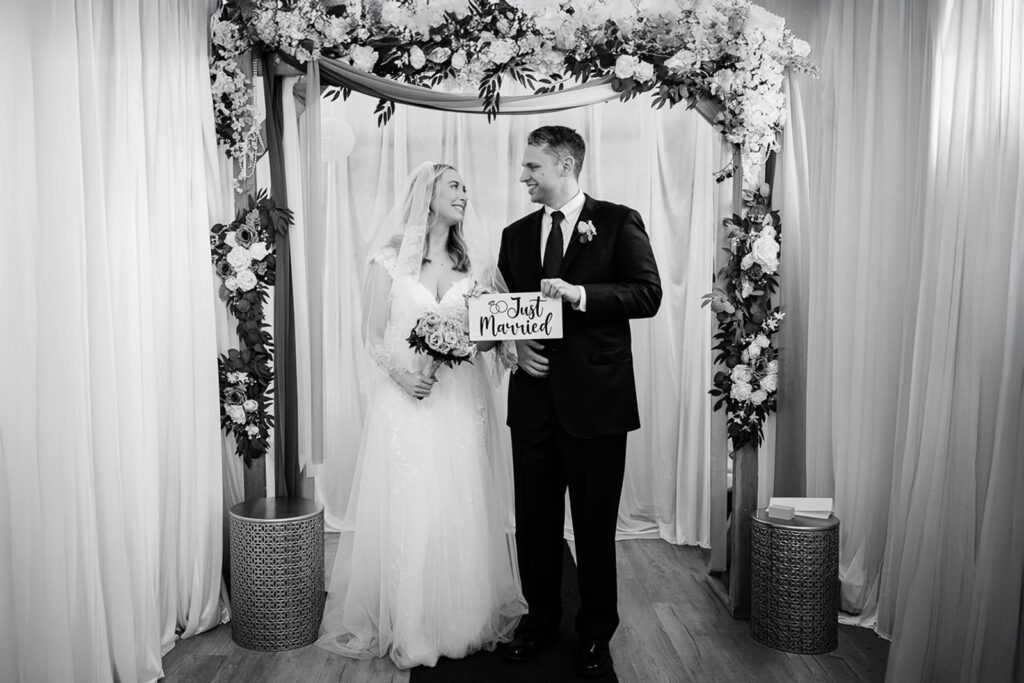 Newlyweds holding Just Married sign under floral wedding arch at The Highlands Mansion and Gardens Fort Washington PA