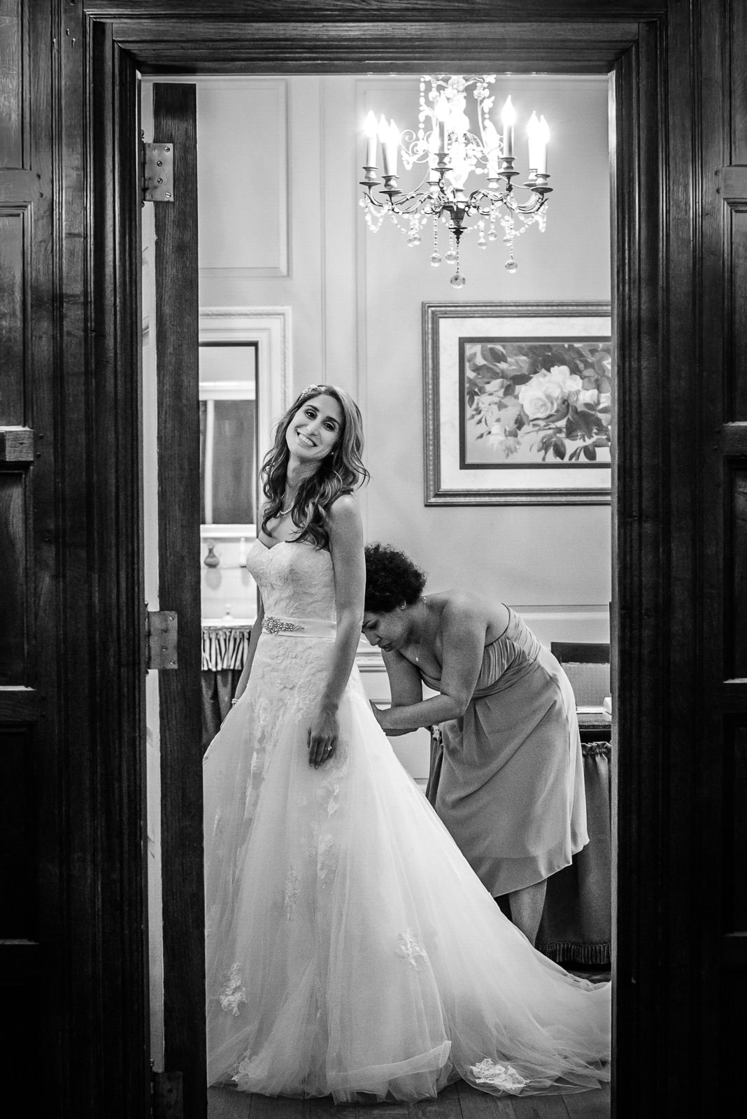 Bride in a strapless lace gown glances back over her shoulder through an elegant doorway while a bridesmaid fastens the back of her dress, captured in black and white by New Jersey wedding photographer Alex Kaplan.