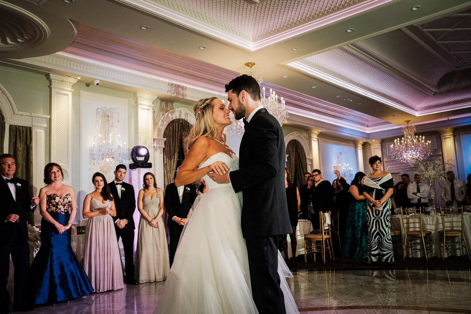 Bride and groom share an intimate first dance moment at Rockleigh Country Club in NJ, surrounded by guests and glowing crystal chandeliers under pink uplighting