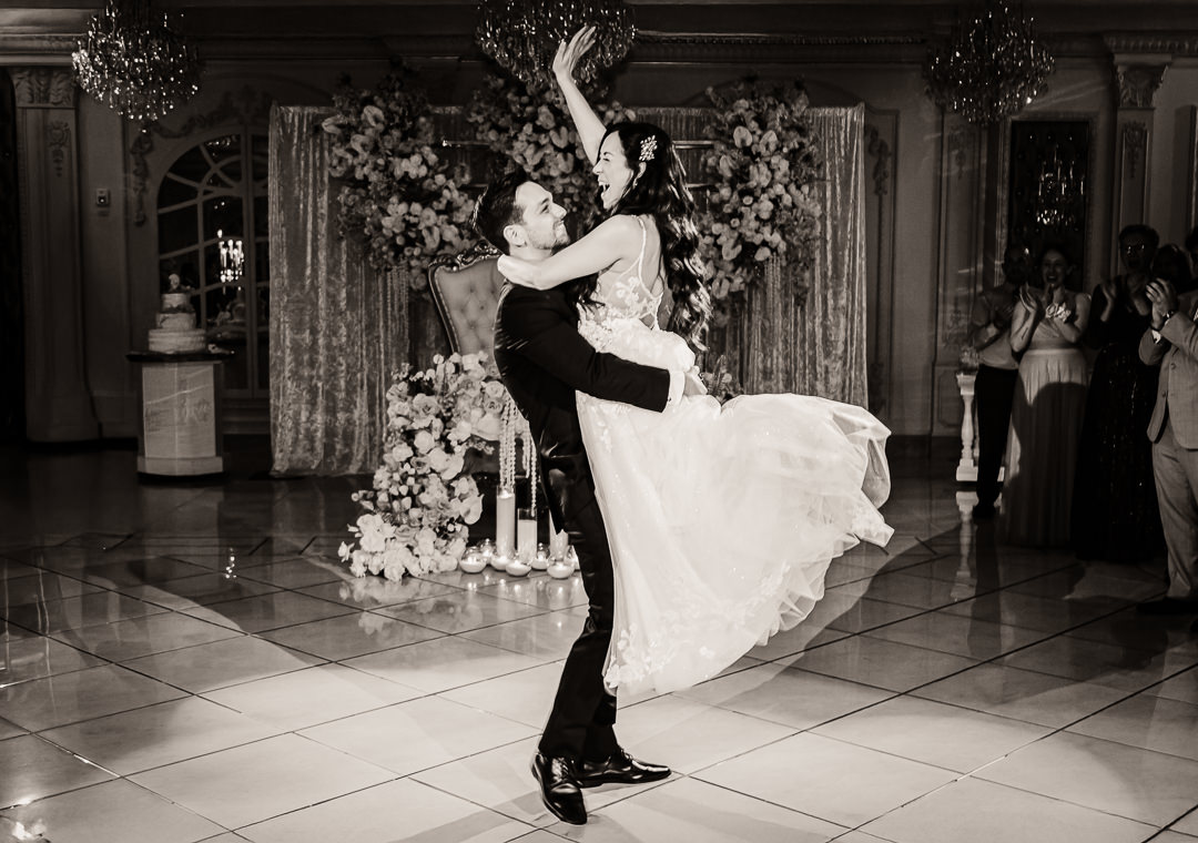 Groom lifting bride joyfully during first dance at luxury New Jersey wedding venue, bride laughing with arm raised, grand floral arrangement in background, black and white