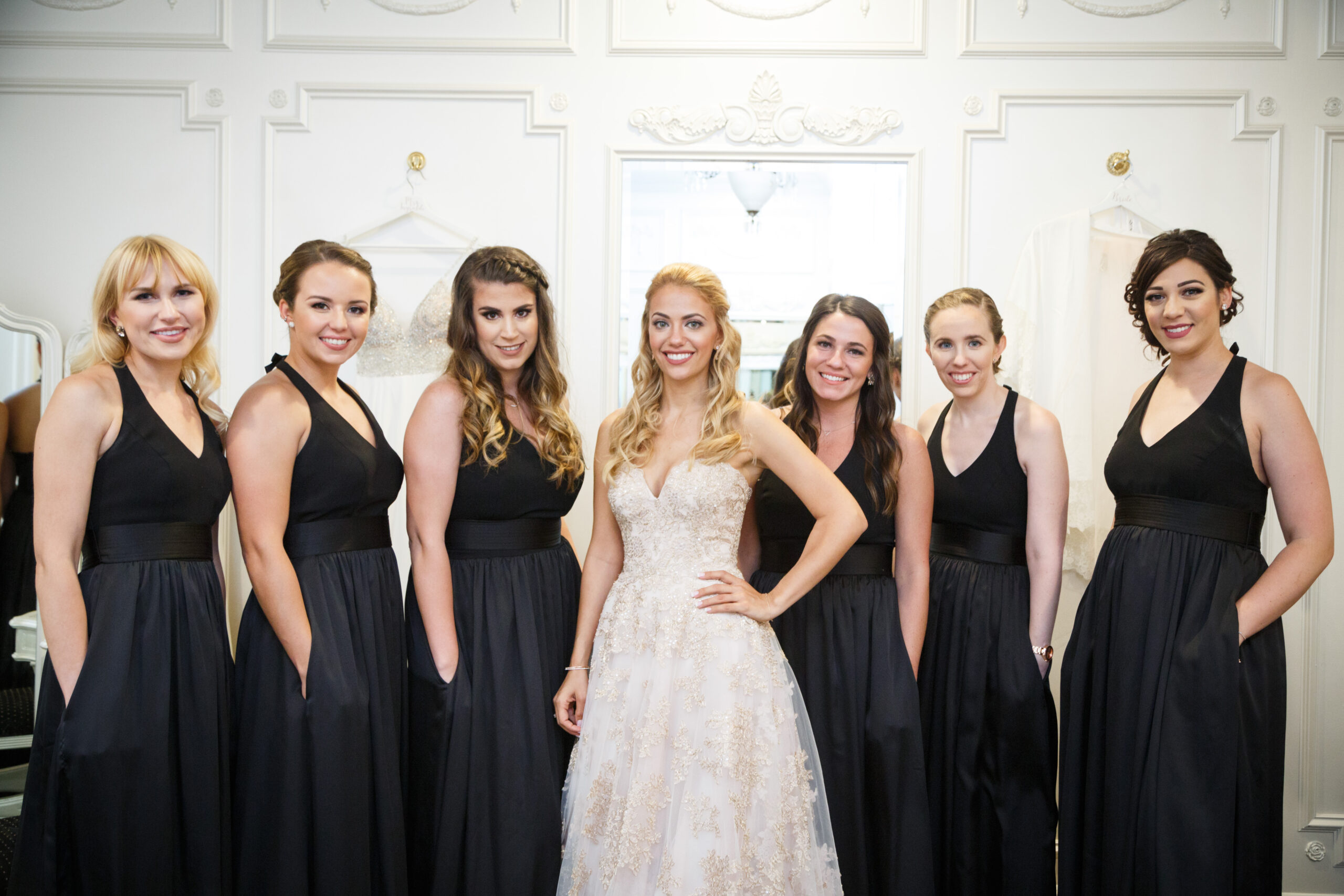 Bride in gold lace ball gown standing with six bridesmaids in black gowns in the Crystal Plaza bridal suite, Livingston NJ, wedding getting ready photography by Alex Kaplan