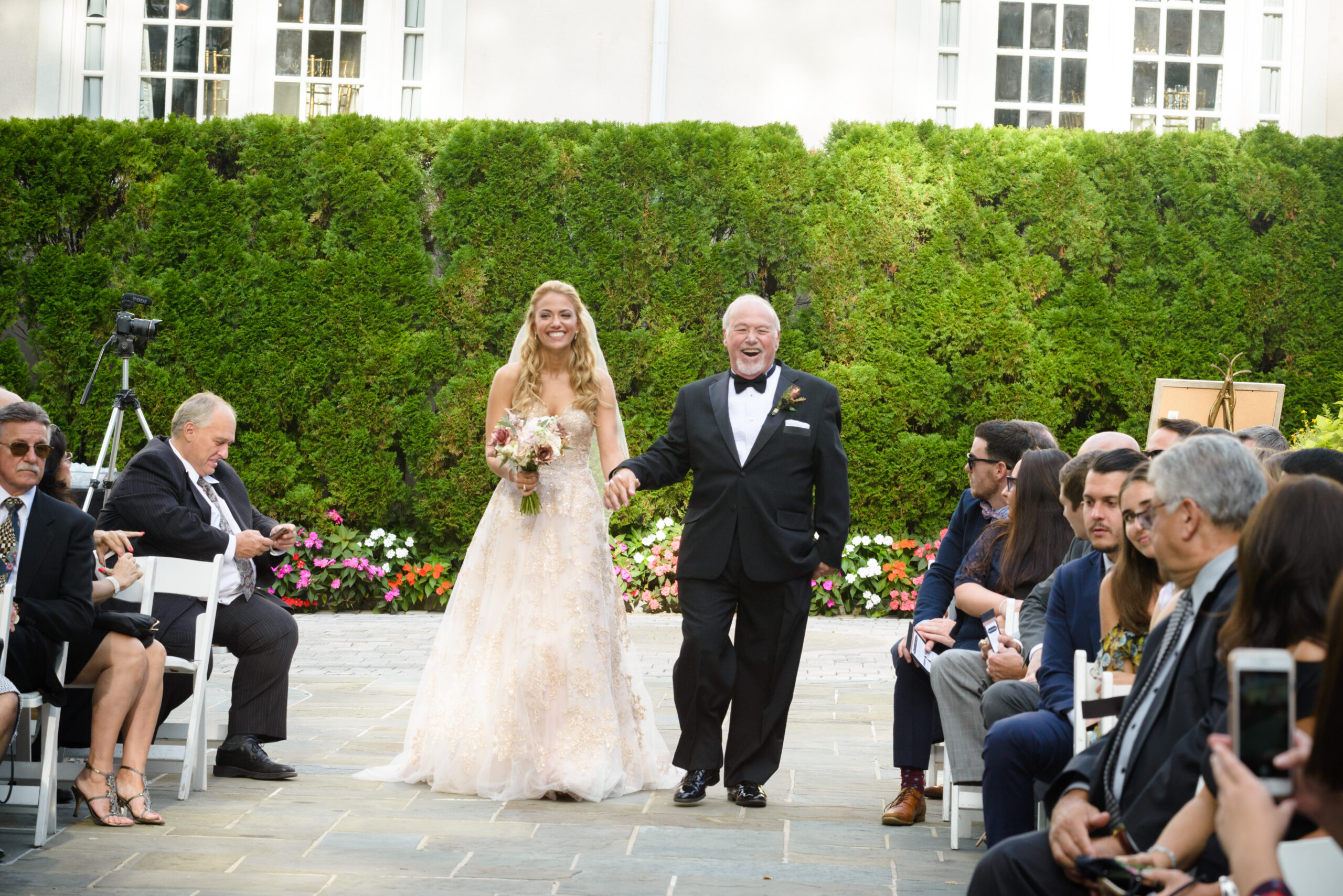 Bride in gold lace ball gown laughing with father while walking down the outdoor Crystal Plaza ceremony aisle on stone patio with garden hedge backdrop, Livingston NJ, wedding ceremony photography by Alex Kaplan