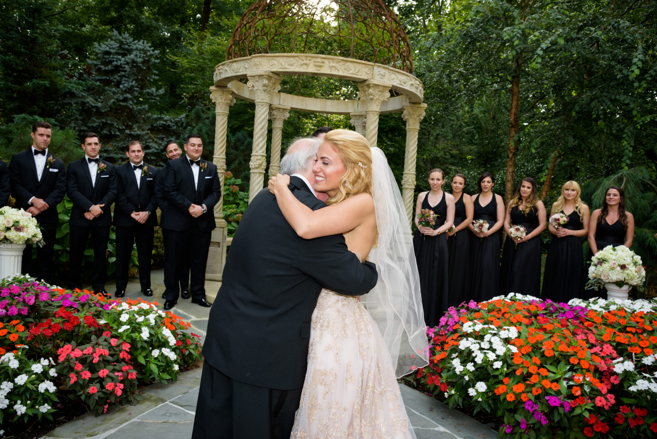 Alt text: Bride in gold lace gown embracing her father at the Crystal Plaza outdoor gazebo ceremony with wedding party and colorful flower gardens in background, Livingston NJ, wedding ceremony photography by Alex Kaplan
