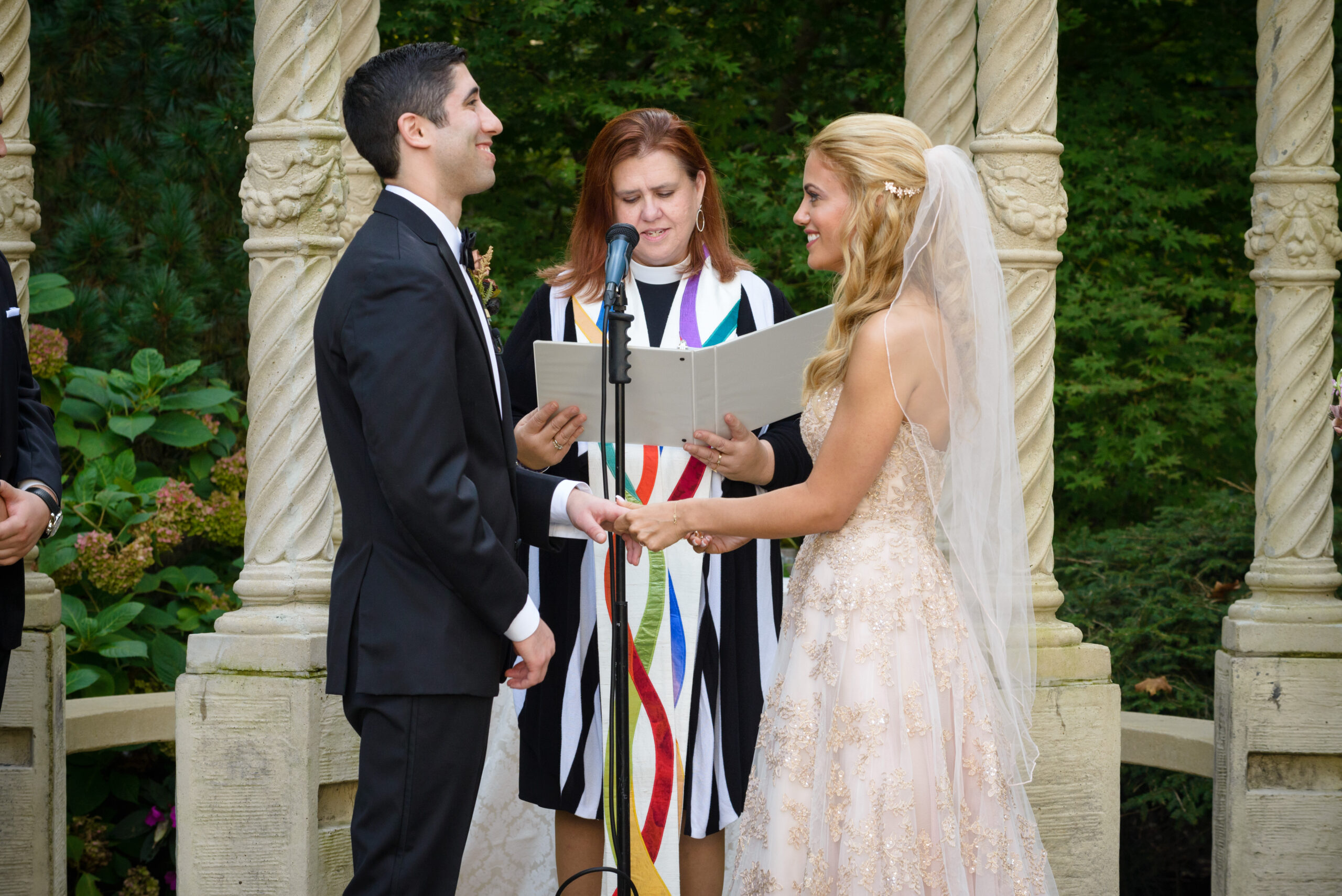 Bride and groom holding hands during vows at the Crystal Plaza outdoor gazebo ceremony with officiant and stone columns, Livingston NJ, outdoor wedding ceremony photography by Alex Kaplan
