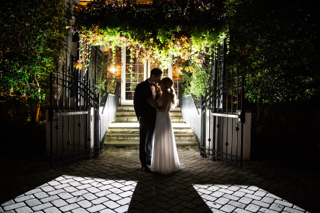 Night portrait of bride and groom silhouetted at Crystal Plaza garden gate with iron fence and steps lit from behind, Livingston NJ, evening wedding portrait by Alex Kaplan