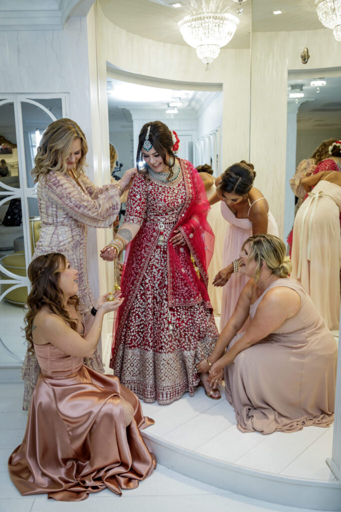 Bridesmaids helping Indian bride into red embroidered lehenga in Crystal Plaza bridal suite before ceremony