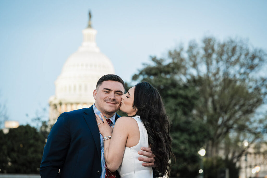 Woman kissing her smiling partner's cheek with the US Capitol dome glowing behind them