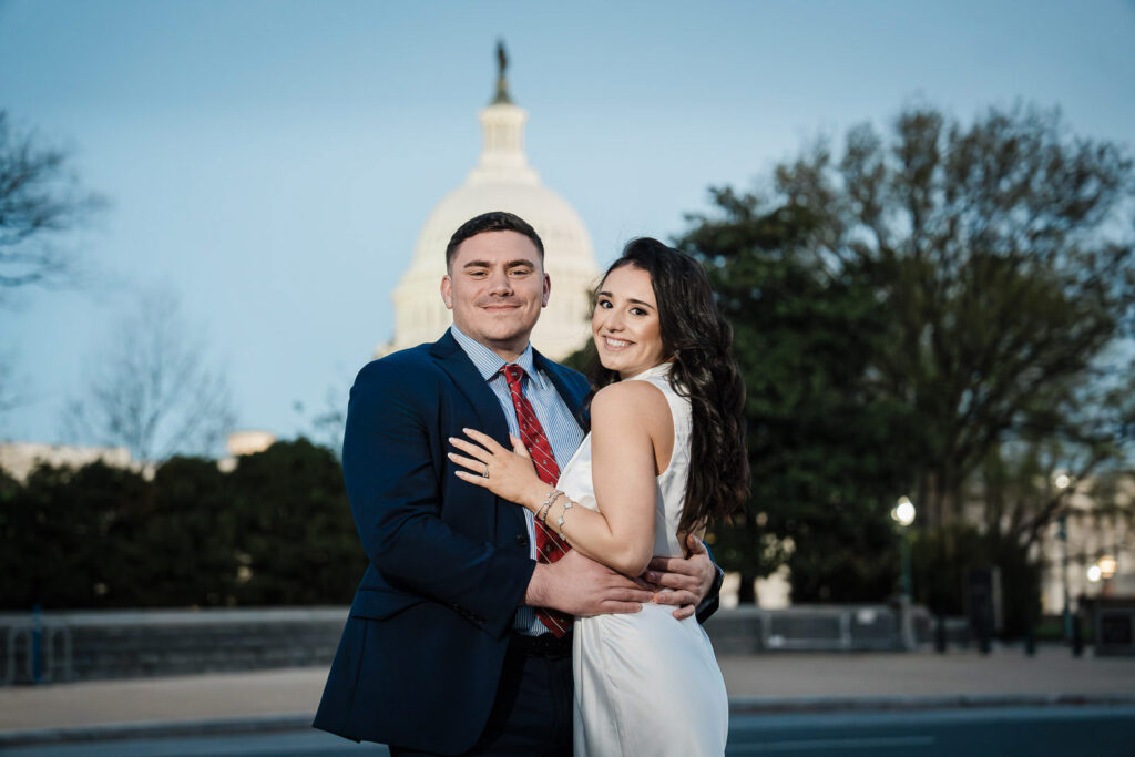 Engaged couple embracing and smiling at the camera with the US Capitol dome behind them