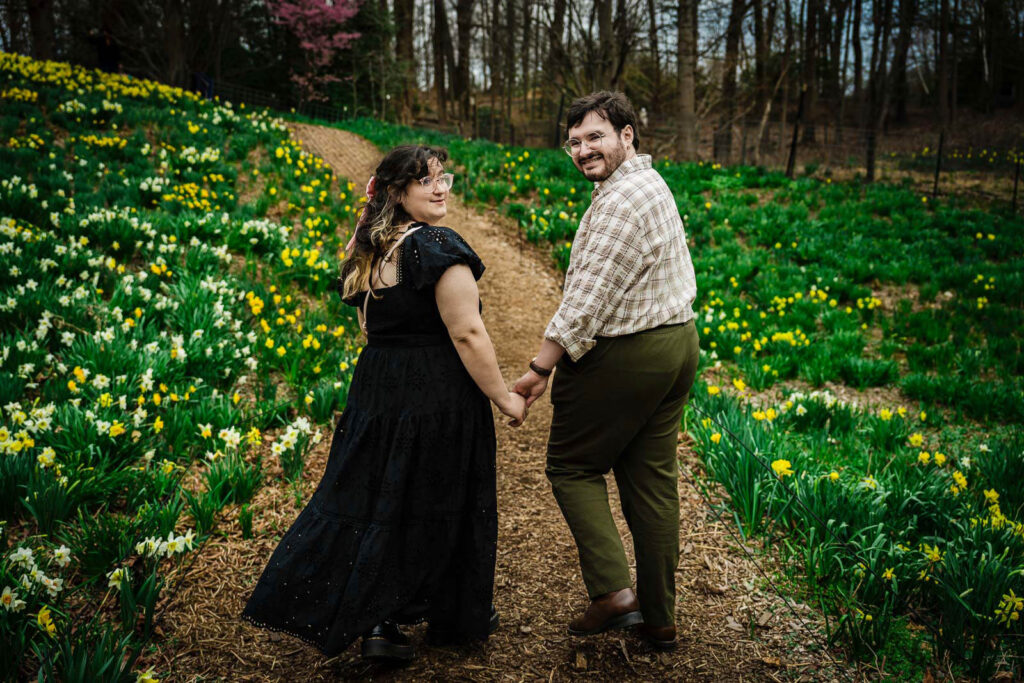 Engaged couple holding hands and walking along daffodil-lined path at Reeves-Reed Arboretum