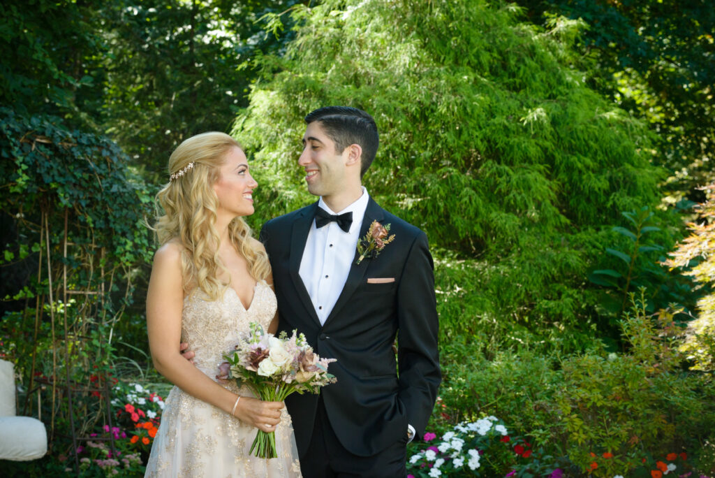 Bride in gold lace gown and groom in black tuxedo standing together in Crystal Plaza garden looking at each other, Livingston NJ, garden wedding portrait by Alex Kaplan