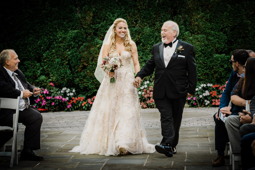Bride in gold lace ball gown laughing with father walking down the outdoor Crystal Plaza ceremony aisle, Livingston NJ, wedding ceremony photography by Alex Kaplan